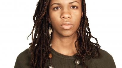 An image of a Black woman chest up, looking straight at the camera. She has long brown braids and is wearing a dark green sweater with a long necklace and dangling earrings. 