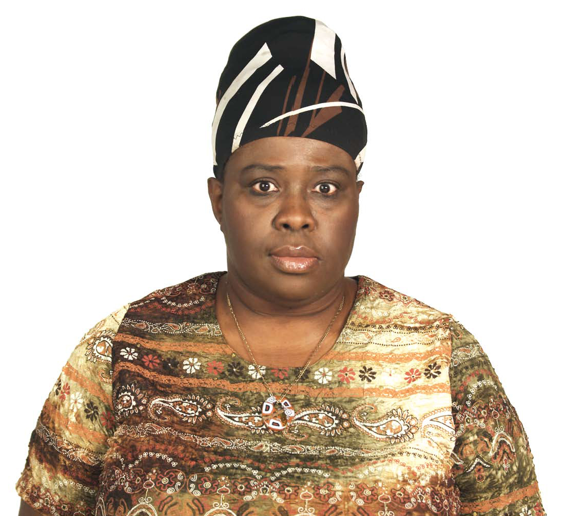 An image of a Black person looking straight at the camera. She is wearing a black, brown, and white patterned head turban and a lighter color patterned shirt with a long necklace.   