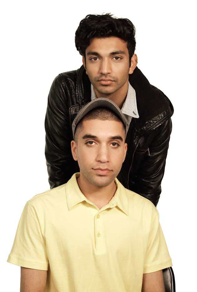 An image of two brown men looking straight at the camera. One of them is wearing a yellow short sleeved shirt and seated, while the other one is standing behind him wearing a black leather jacket.  