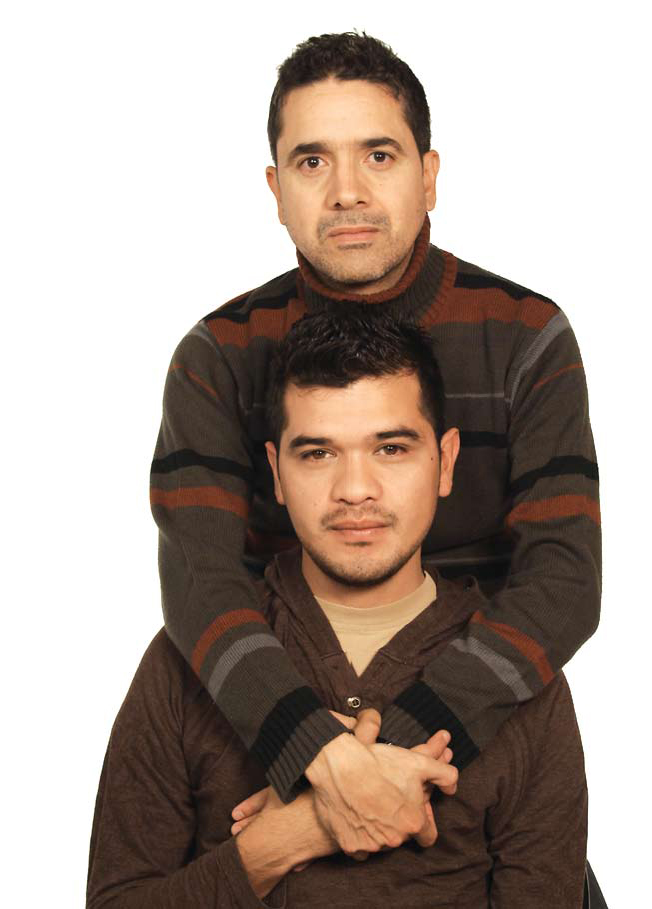 An image of two brown man looking straight at the camera. One of them is appears to be sitting, the other one is standing behind, holding each other's hands. They both wear brown sweaters.