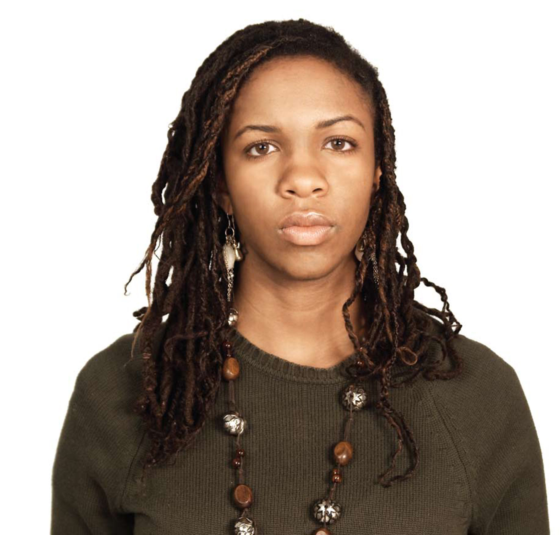 An image of a Black woman chest up, looking straight at the camera. She has long brown braids and is wearing a dark green sweater with a long necklace and dangling earrings. 