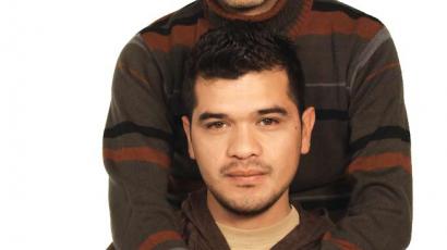 An image of two brown man looking straight at the camera. One of them is appears to be sitting, the other one is standing behind, holding each other's hands. They both wear brown sweaters.